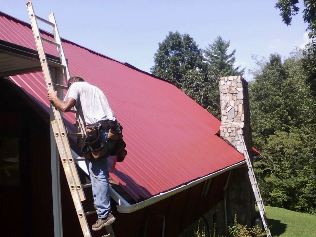 Roofer on a ladder beside a red metal roof and stone chimney — Soffit & Fascia Repair by Roofing Brothers in Bensenville