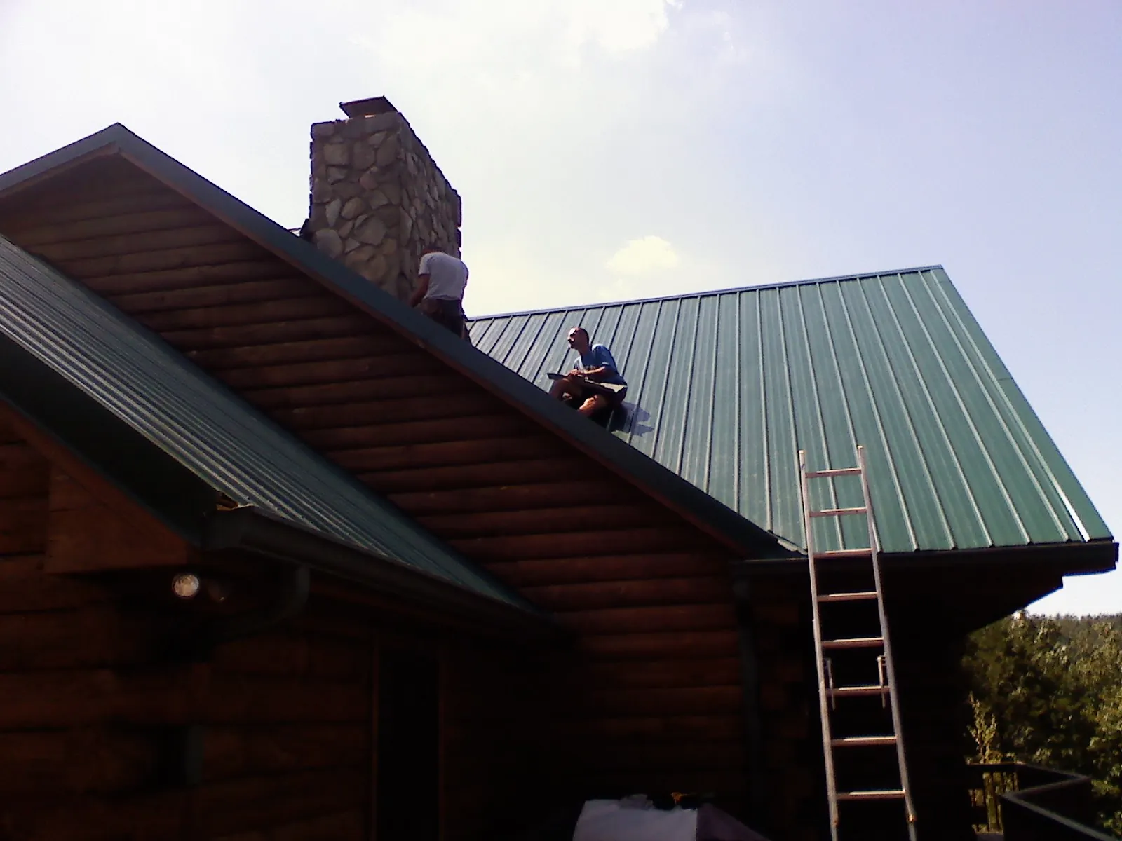 Roofers installing green metal panels beside a stone chimney on a log home in Bensenville